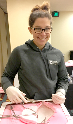 Sara Rummeholff wearing a hoodie, and surgical gloves. She's smiling and practicing surgery on a foam dummy.