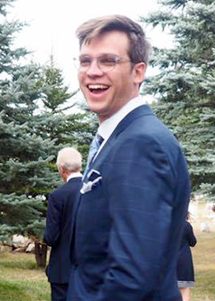 Cameron wearing a suit outside around some trees. He's smiling.